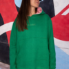 Sudadera verde con capucha y bolsillo frontal, estilo casual, combinada con jeans frente a un mural colorido.