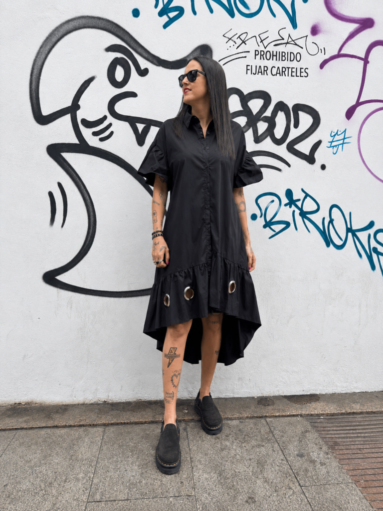 Vestido camisero negro oversize para mujer con corte asimétrico, volantes y detalles metálicos decorativos.