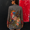 Mujer vistiendo la Blazer Guacamayo con estampado floral multicolor y gran guacamayo tropical sobre tejido de cuadro príncipe de Gales enseñando la parte trasera