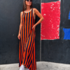 Mujer posando con vestido largo de rayas verticales naranja y azul marino, cuello redondo beige y zapatillas blancas sobre un fondo oscuro.