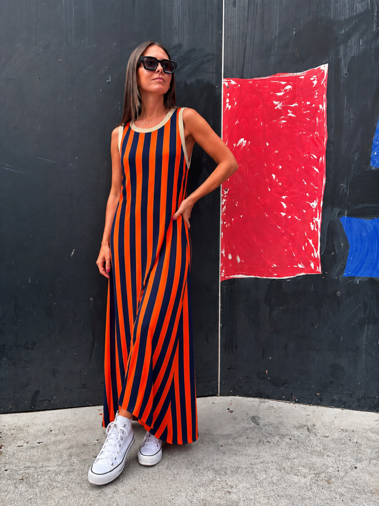 Mujer posando con vestido largo de rayas verticales naranja y azul marino, cuello redondo beige y zapatillas blancas sobre un fondo oscuro.