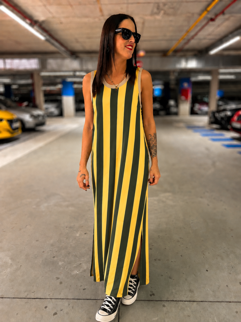 Mujer luciendo un vestido maxi de rayas verticales amarillo y verde oliva, sin mangas y con zapatillas negras, en un entorno urbano.