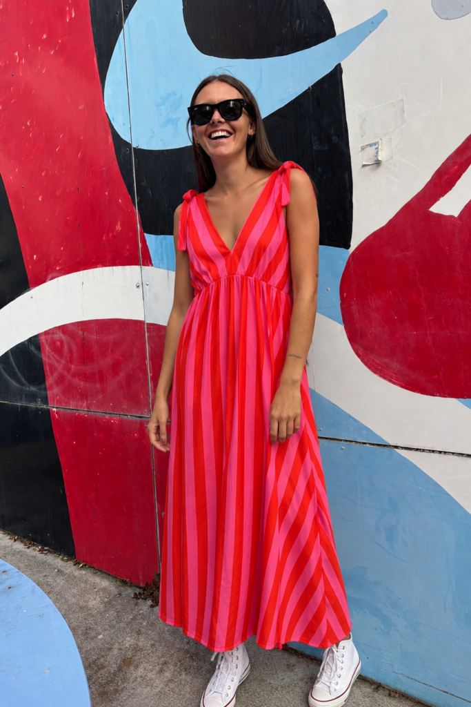 Mujer luciendo el Vestido Flamingo, un vestido maxi de rayas rosa y rojo con escote en V y tirantes con lazadas.
